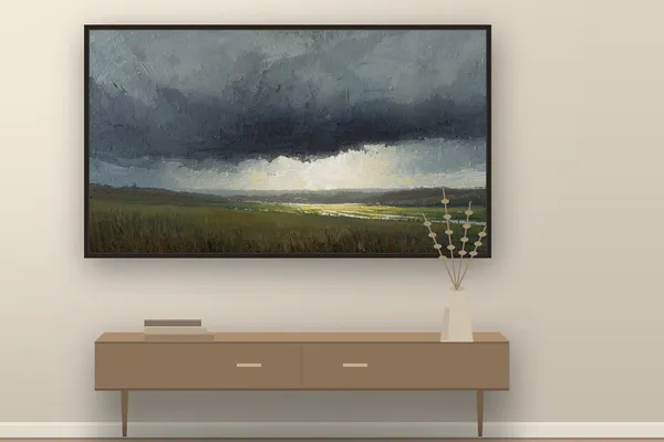 A Samsung Frame TV on a warm cream wall showing a quiet painterly landscape of a low storm over green fields, above a wooden console with a pale vase