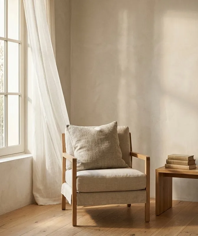A cream linen armchair with a folded pillow, tucked into a sunlit corner beside sheer curtains, with a small stack of books resting on a wooden side table.