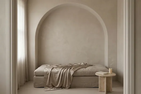 A linen daybed with a rumpled matte champagne satin throw pulled halfway back inside a plaster arch alcove, with a small travertine side table holding a closed cream journal and a candle with a thin wisp of smoke rising