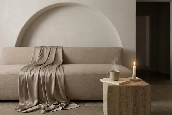 A linen sofa under a plaster arch with sparse shelves above, champagne satin draped over the arm, a steaming mug and closed notebook on a travertine block, and one candle in soft ivory light