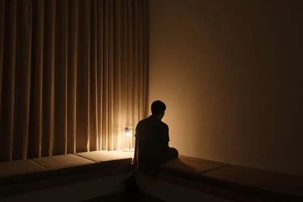 A dark room at night: a figure sits on a low platform with their back to us, a small lamp glowing against a floor-length curtain
