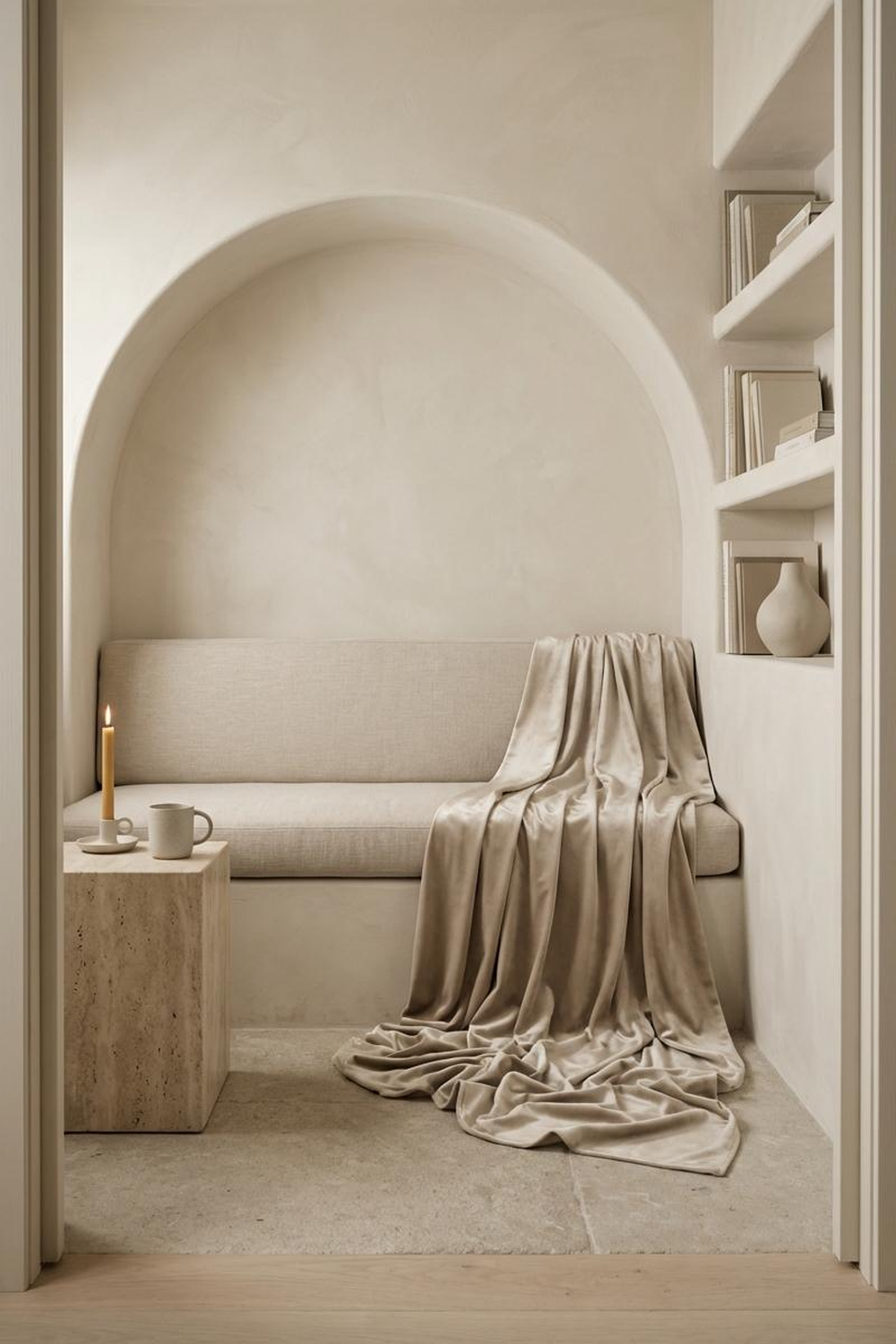 An arched alcove with linen banquette, silk drape pooling on stone floor, travertine side table with candle and mug, bookshelves with cream books and ceramic vase
