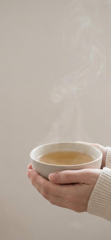 two hands cradling a pale bowl of chamomile tea, steam rising into empty warm-grey space
